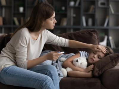 Mulher está aferindo a temperatura da febre filha, que está deitada no sofá abraçando um ursinho branco.
