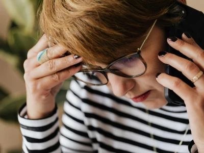 Mulher de óculos com expressão de dor, segurando a cabeça com uma mão enquanto fala ao telefone, ilustrando o impacto da cefaleia no dia a dia e a importância de buscar informação.