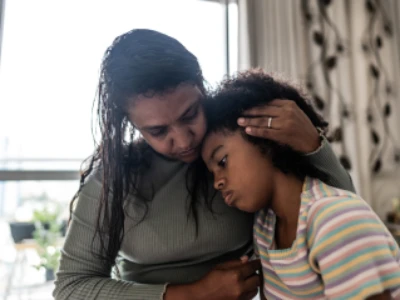 Mãe abraça sua filha, oferecendo a ela um conforto ao aninhar a cabeça dela em seu próprio peito.