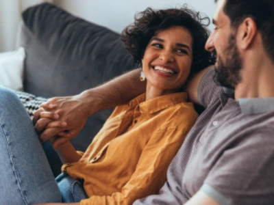 Casal sentado em sofá, olhando carinhosamente um para o outro e sorrindo.