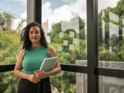 Mulher segurando uma pasta de trabalho diante de uma janela. Ao fundo é possível observar uma área verde. Ela tem uma expressão serena. 