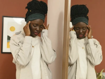 Mulher com faixa de cabelo verde, vestindo uma camisa cinza-clara, está de pé em um quarto, segurando as têmporas e a testa com as duas mãos, refletida em um espelho ao fundo, transmitindo uma sensação de dor de cabeça ou sinusite.