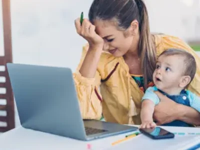 Mãe sentada em frente a um laptop, está com semblante de dor, apoiando a cabeça em uma mão e segurando o bebê com a outra.