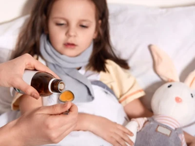 Uma mãe administra dipirona em gotas para sua filha deitada na cama, que usa um cachecol e tem um coelho de pelúcia ao lado, destacando o desafio da dosagem com colheres.