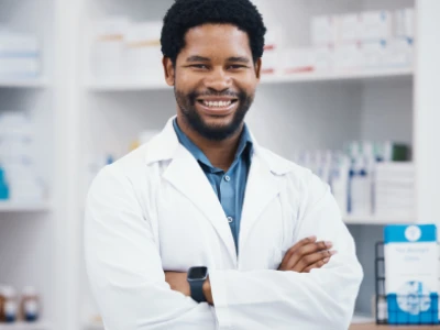 Farmacêutico em drogaria, de braços cruzados, sorrindo fazendo pose profissional para a foto.