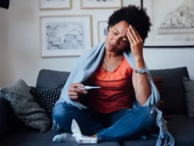 Mulher sentada no sofá, medindo temperatura do corpo com termômetro e com a mão na cabeça com fisionomia de dor