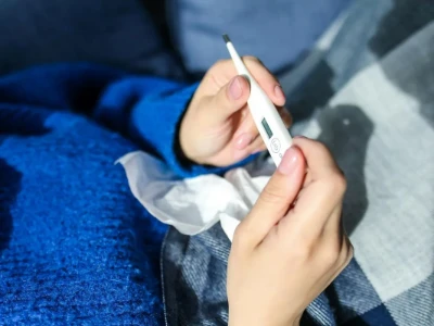 Close-up das mãos de uma pessoa que, agasalhada com um suéter azul e um cobertor, segura um termômetro digital para verificar a temperatura. A imagem foca no ato de medir a febre durante um quadro de doença.
