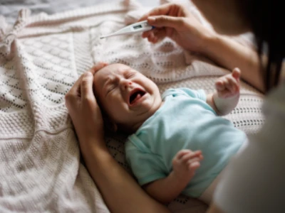 Bebê deitado chorando é confortado pela mãe, que acaba de aferir a sua temperatura com um termômetro.