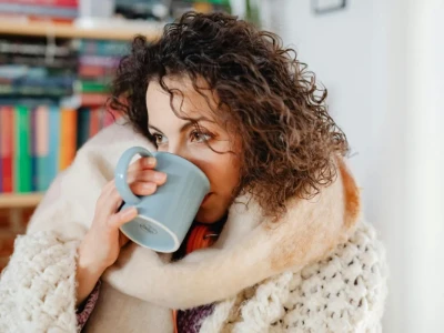 Uma mulher agasalhada, com um lenço, bebe um chá quente de uma caneca, ilustrando o conforto e o alívio que bebidas quentes podem trazer durante um resfriado