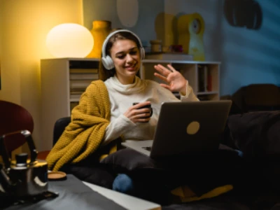 Mulher com fones de ouvido, sentada em sua sala de estar, está coberta por manta, segurando xícara de chá enquanto sorri para a câmera do laptop.