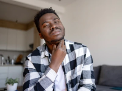 Homem negro vestindo camisa xadrez, com os olhos fechados e a mão no pescoço, demonstrando expressão de dor e desconforto na garganta.