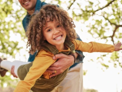 Pai sorridente segurando sua filha pequena nos braços, levantando-a como se ela estivesse voando, em um parque com árvores ao fundo. A imagem transmite felicidade e saúde após o tratamento.