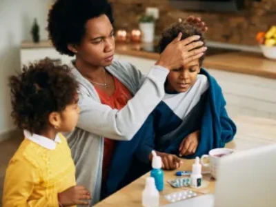 Uma mãe verifica a temperatura do filho colocando a mão na testa dele, enquanto uma criança menor observa; frascos de remédio estão sobre a mesa à frente.