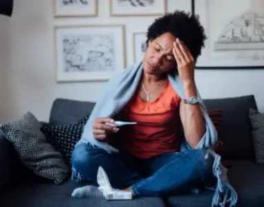 Mulher sentada no sofá, medindo temperatura do corpo com termômetro e com a mão na cabeça com fisionomia de dor