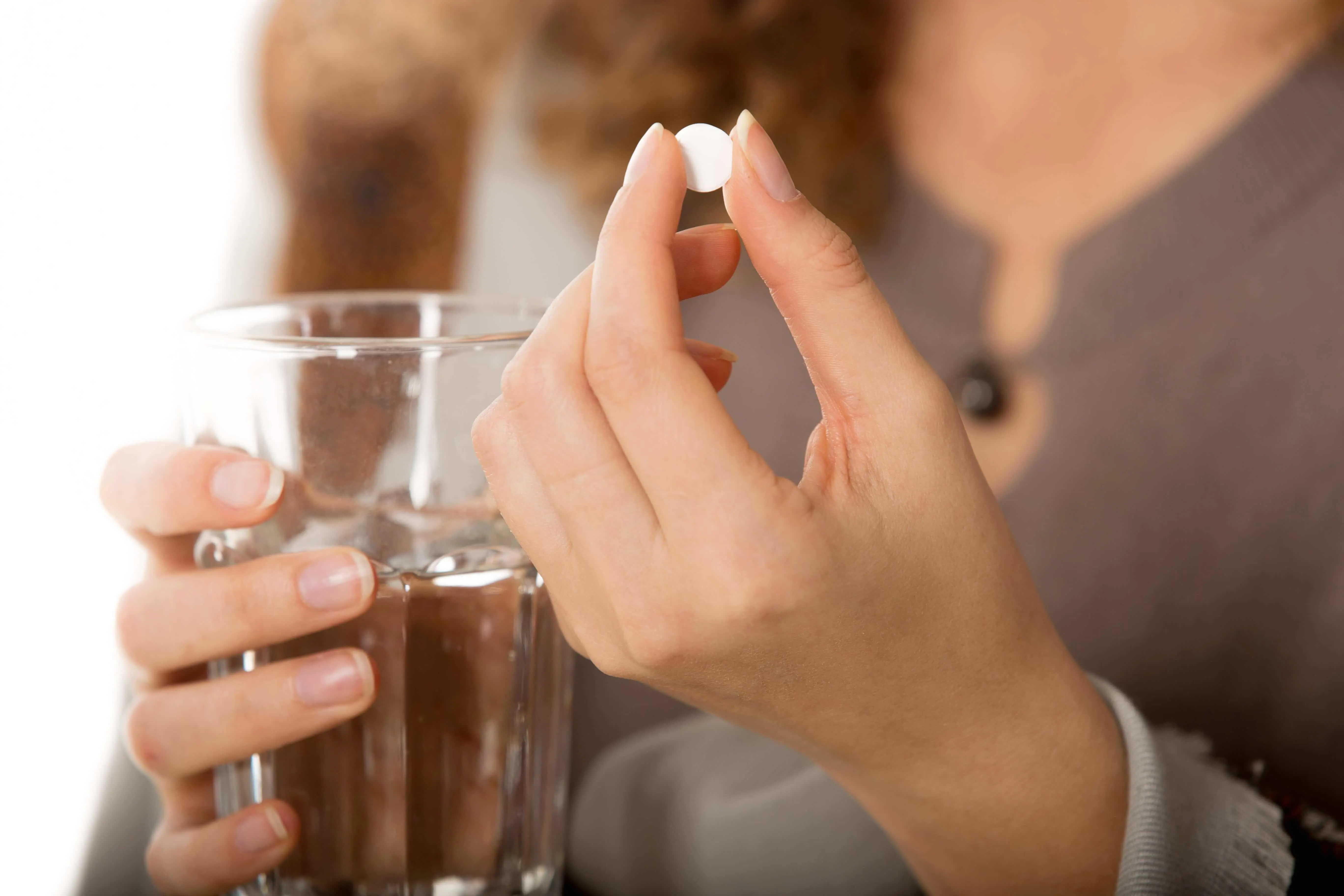 Mulher segurando um copo com água na mão direita e entre os dedos da mão esquerda um comprimido de remédio.