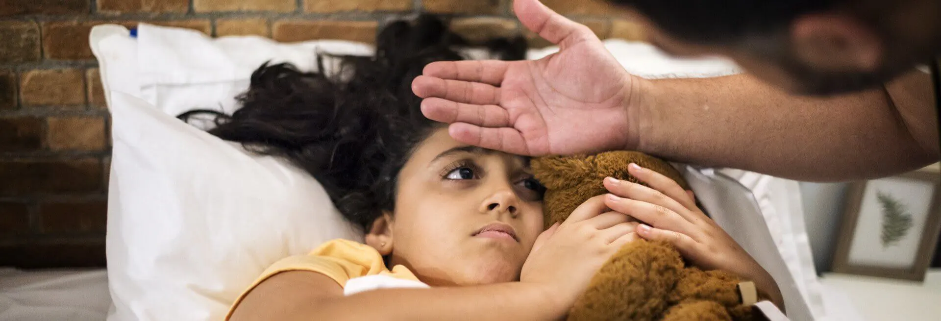 Criança deitada abraçada com um ursinho enquanto adulto verifica se está com febre com a mão sobre a testa da criança