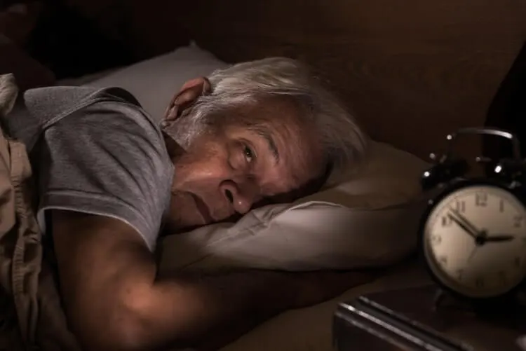 Homem de idade, deitado na cama de bruços de olhos abertos, aparentemente cansado e relógio de cabeçeira marcando 2:53.