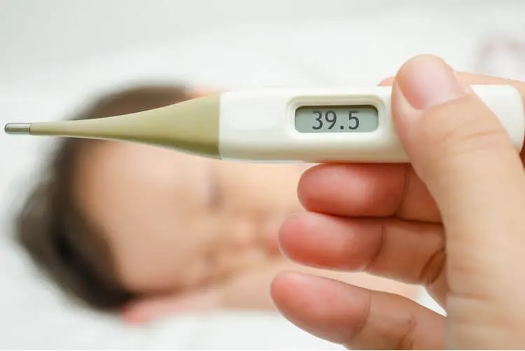 Mão segurando um termômetro marcando 39.5° e ao fundo, um bebê dormindo