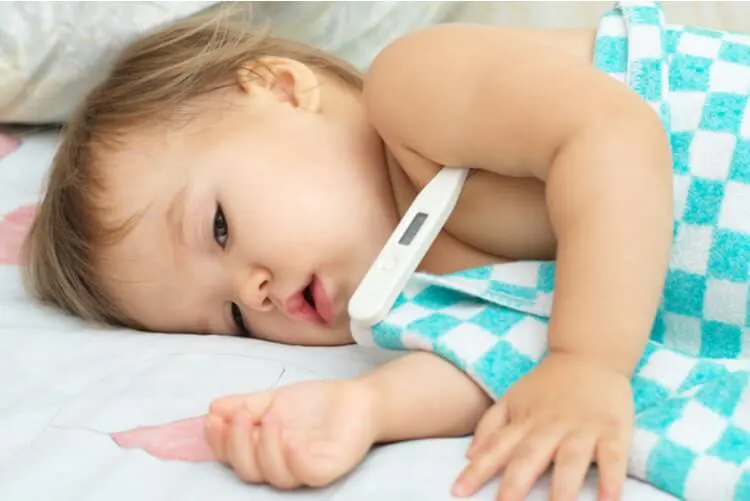 Bebê deitado de lado na cama, com termômetro em baixo do braço, aparentemente doente.