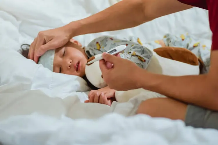 Criança deitada de lado na cama. Pessoa conferindo a febre, com termômetro na mãe e segurando uma toalha na testa da criança.