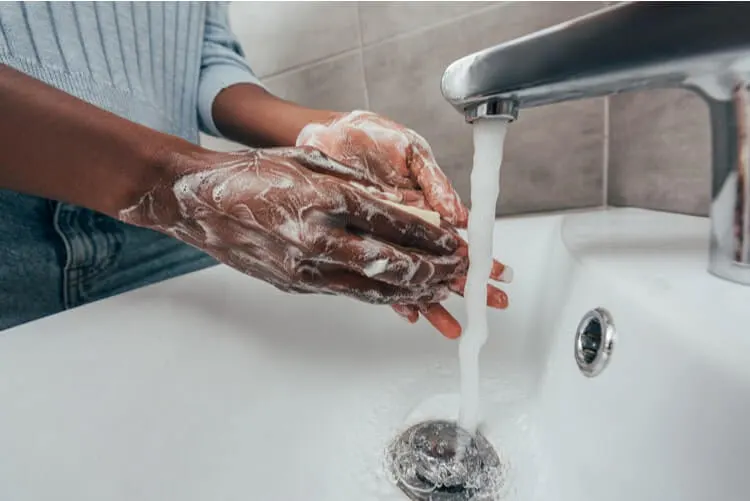 Pessoa preta lavando as mãos em uma pia. Mãos ensaboadas e torneira aberta.