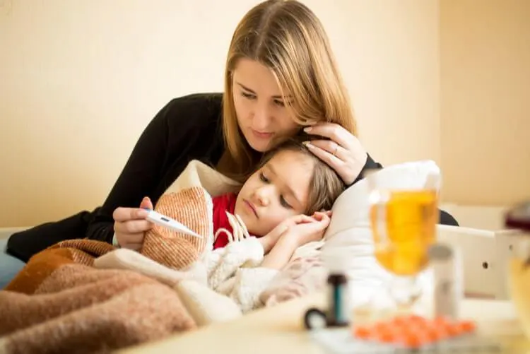 Mulher cuidando de uma criança, aparentemente doente, que está deitada na cama e ambas estão olhando para o termômetro na mão da mulher.
