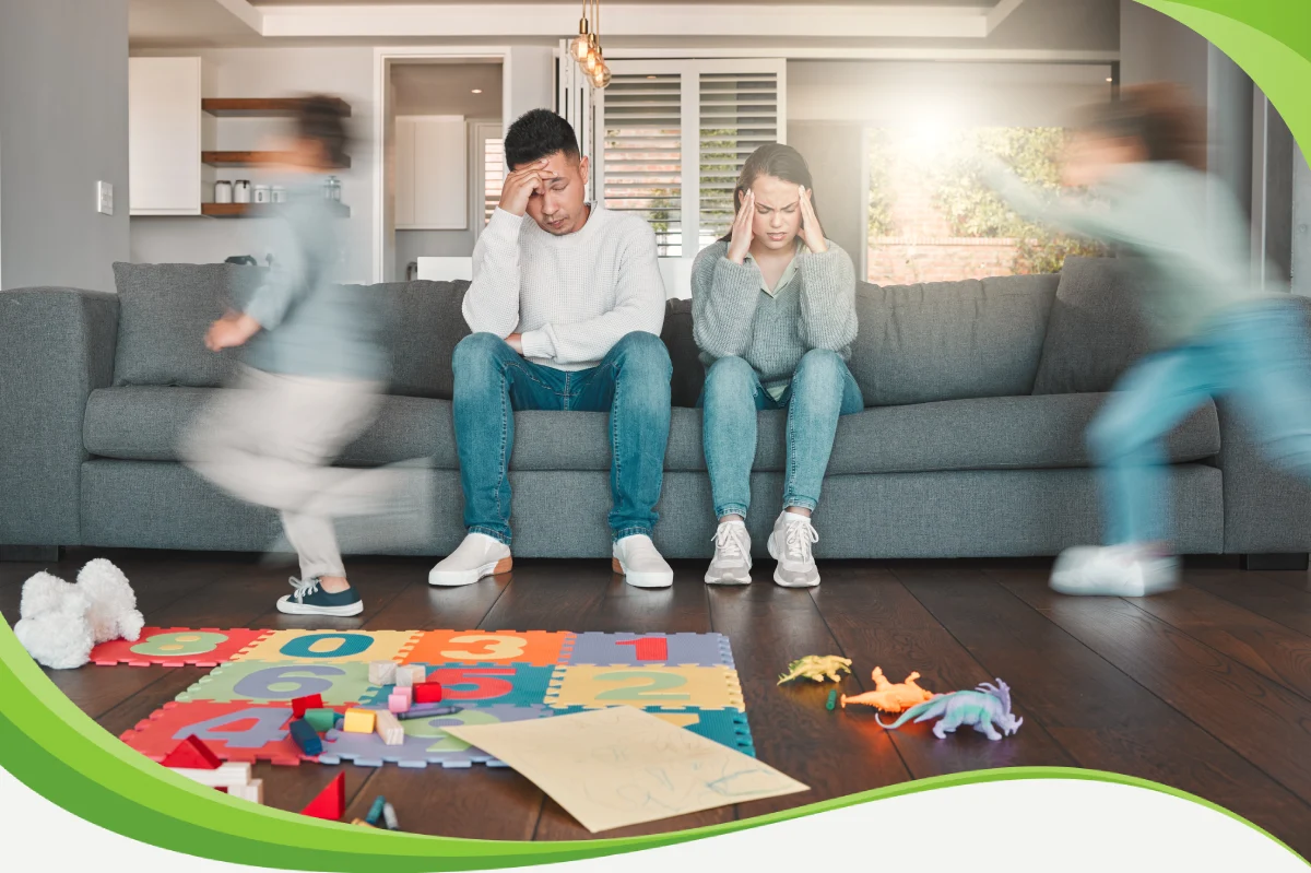 Pais sentados no sofá, com as mãos nas têmporas, parecem cansados enquanto seus filhos correm pela sala