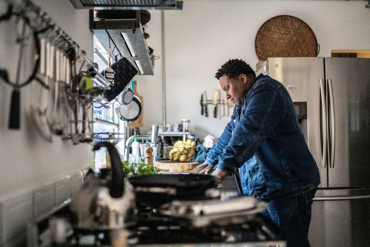 Homem está em pé em sua cozinha, com a cabeça baixa, ombros encolhidos e mãos sobre a pia.