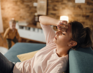 Mãe sentada no sofá com uma mão sobre a testa. Ela parece cansada e com dor.