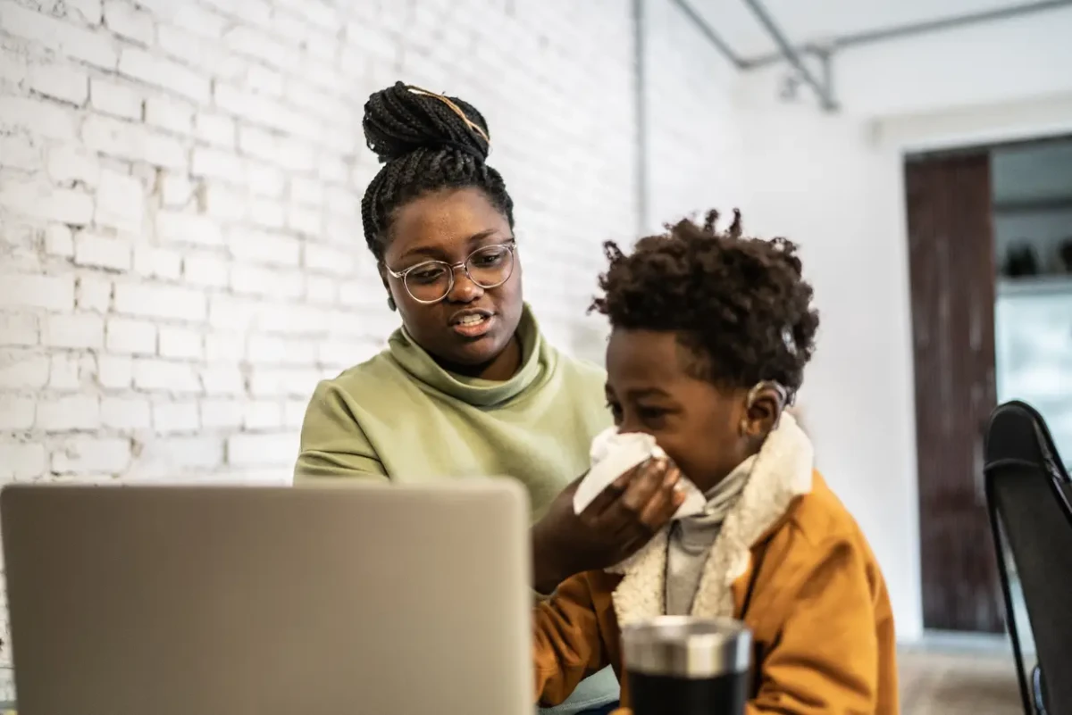 Mãe e filho olhando para a tela de um notebook; a criança segura um lenço de papel próximo ao nariz, ilustrando o cuidado com a coriza e a busca por orientações médicas ou telemedicina.