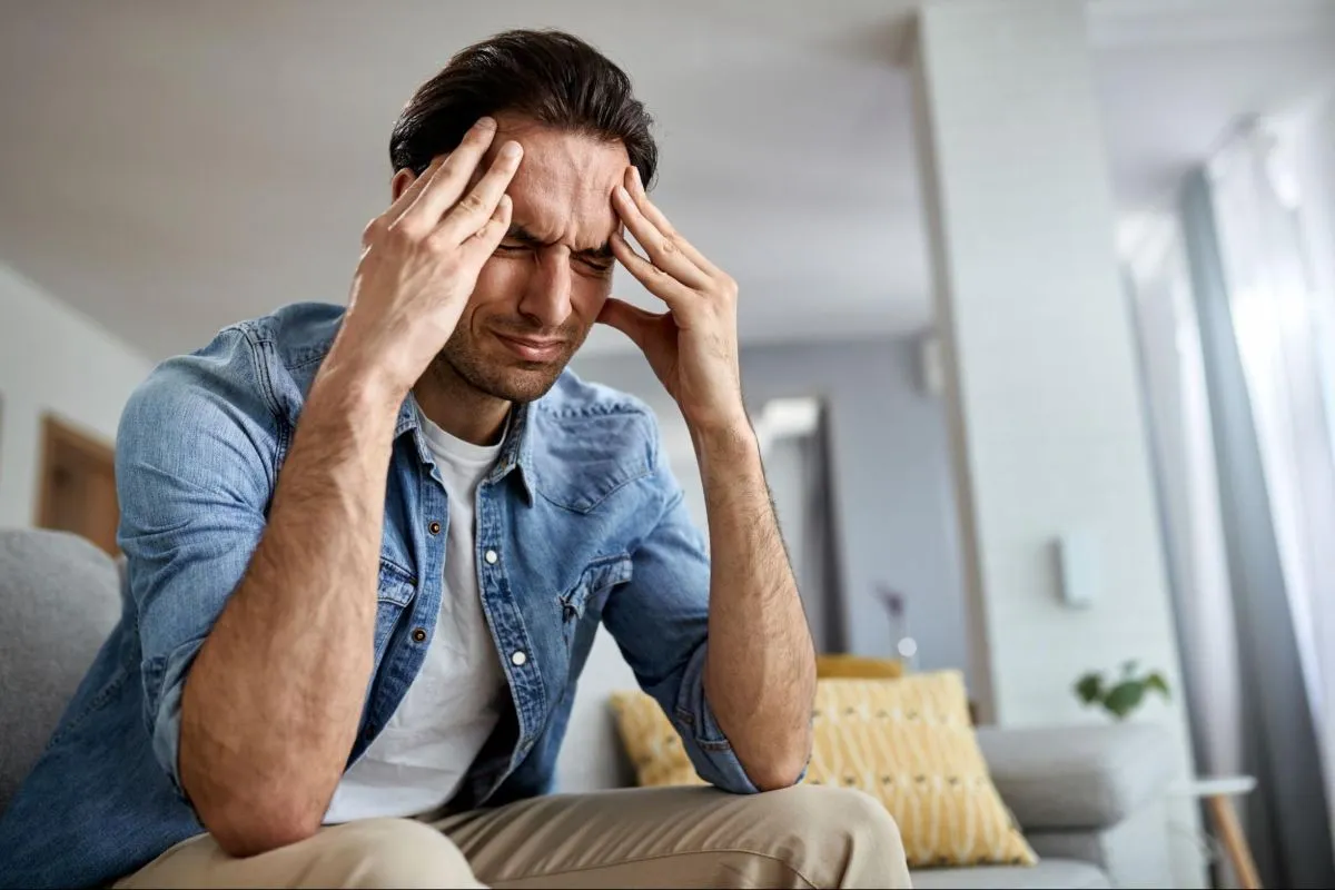 Homem sentado em um sofá, vestindo camisa jeans, com os olhos fechados e os dedos pressionando as têmporas, demonstrando forte dor de cabeça ou enxaqueca.