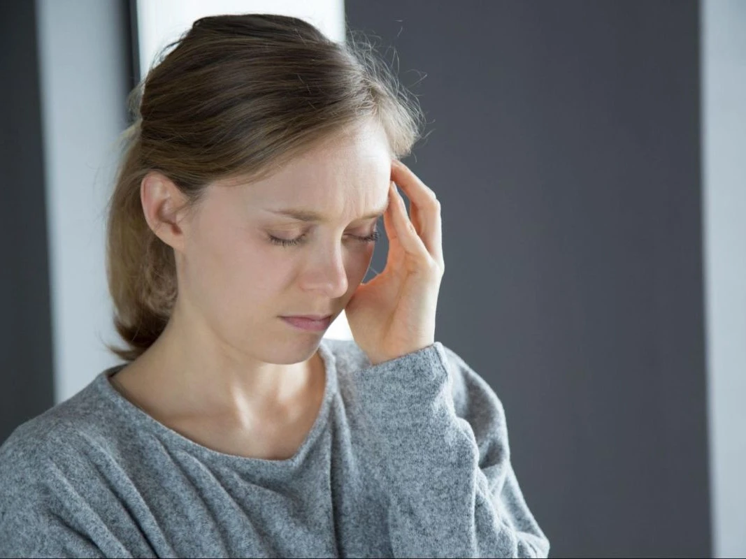 Close de uma mulher com blusa cinza e olhos fechados, massageando a têmpora esquerda com a mão para tentar aliviar uma dor de cabeça ou enxaqueca.