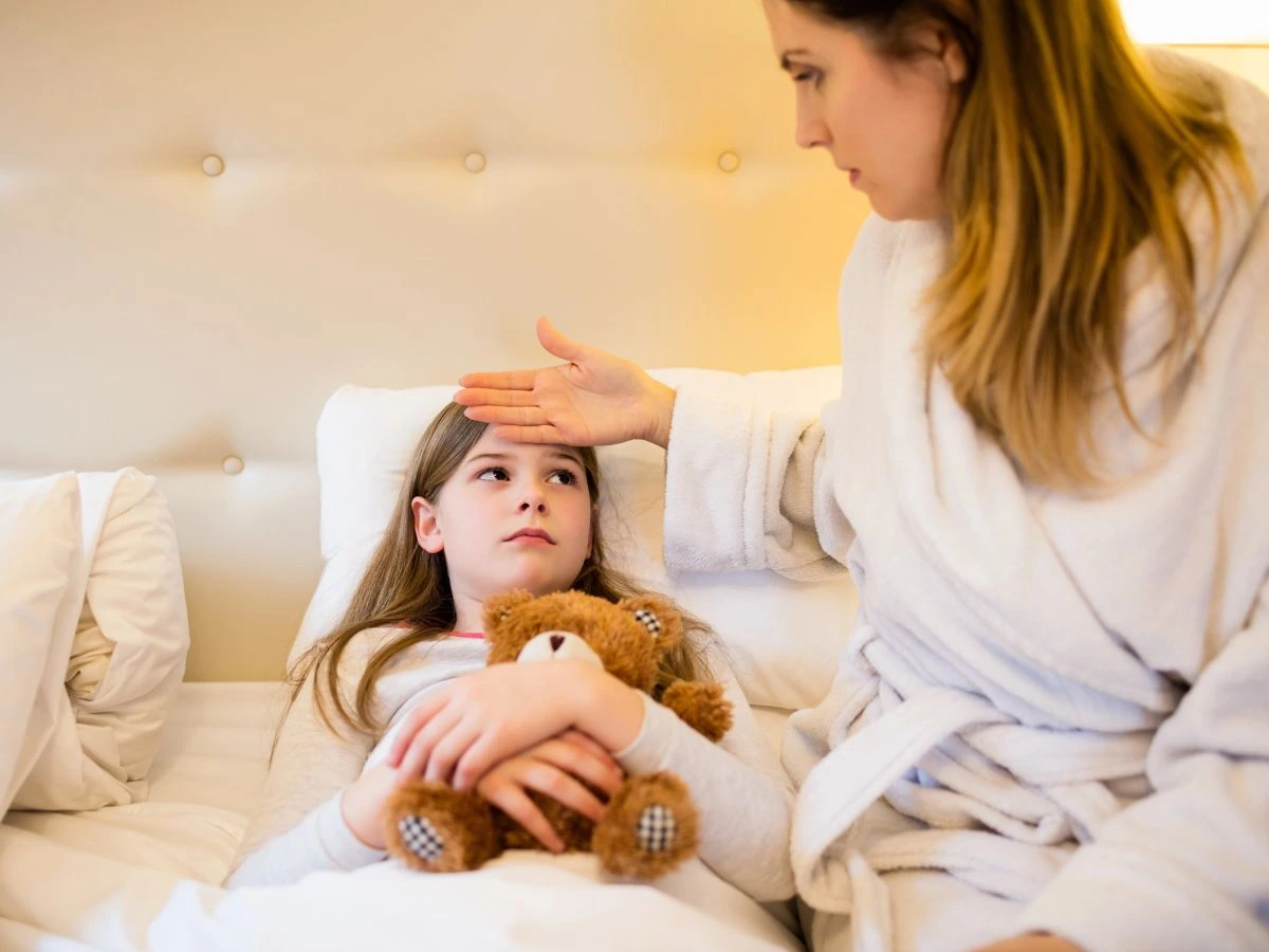 Mãe preocupada mede a temperatura da filha colocando a mão na testa da menina, que está deitada na cama, demonstrando um dos sinais de febre.