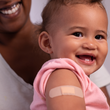 Bebê sorridente, no colo da mãe, com um adesivo no braço logo depois de tomar ser vacinada