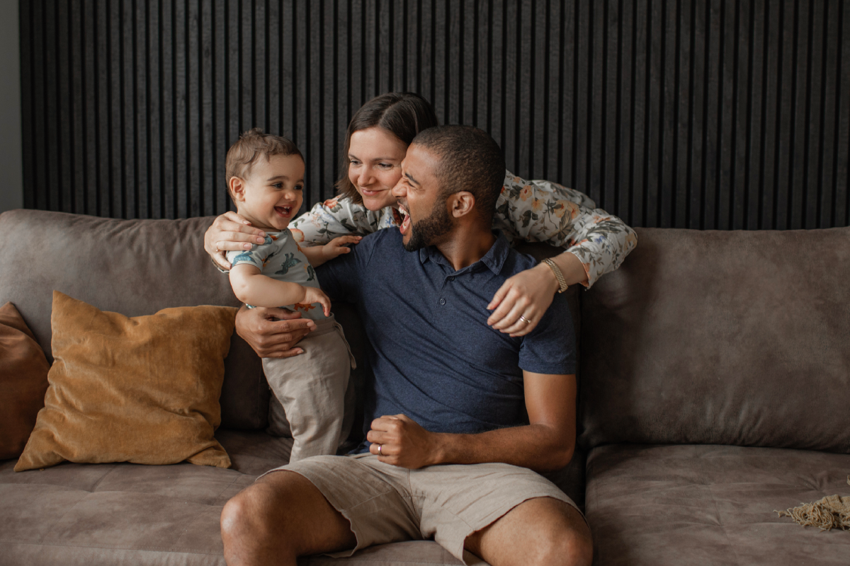 Família feliz no sofá. O pai está sentado, segurando o bebê que está de pé ao seu lado. A mãe vem por trás e abraça ambos.