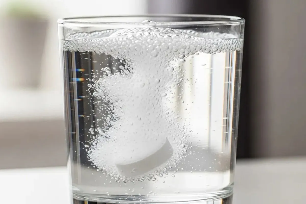 Close-up de um comprimido efervescente branco dissolvendo em um copo de vidro com água, criando muitas bolhas.