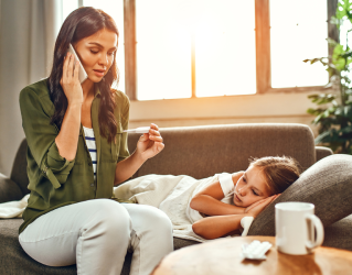 Criança deitada no sofá, enquanto sua mãe está sentada ao seu lado e parece falar ao telefone preocupada.