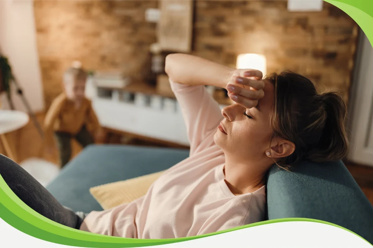 Mãe sentada no sofá com uma mão sobre a testa. Ela parece cansada e com dor.anheiro no sofá, abraçada nele. Ele está tocando as têmporas, com feições de dor.