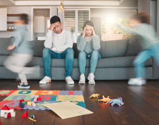 Pais sentados no sofá, com as mãos nas têmporas, parecem cansados enquanto seus filhos correm pela sala