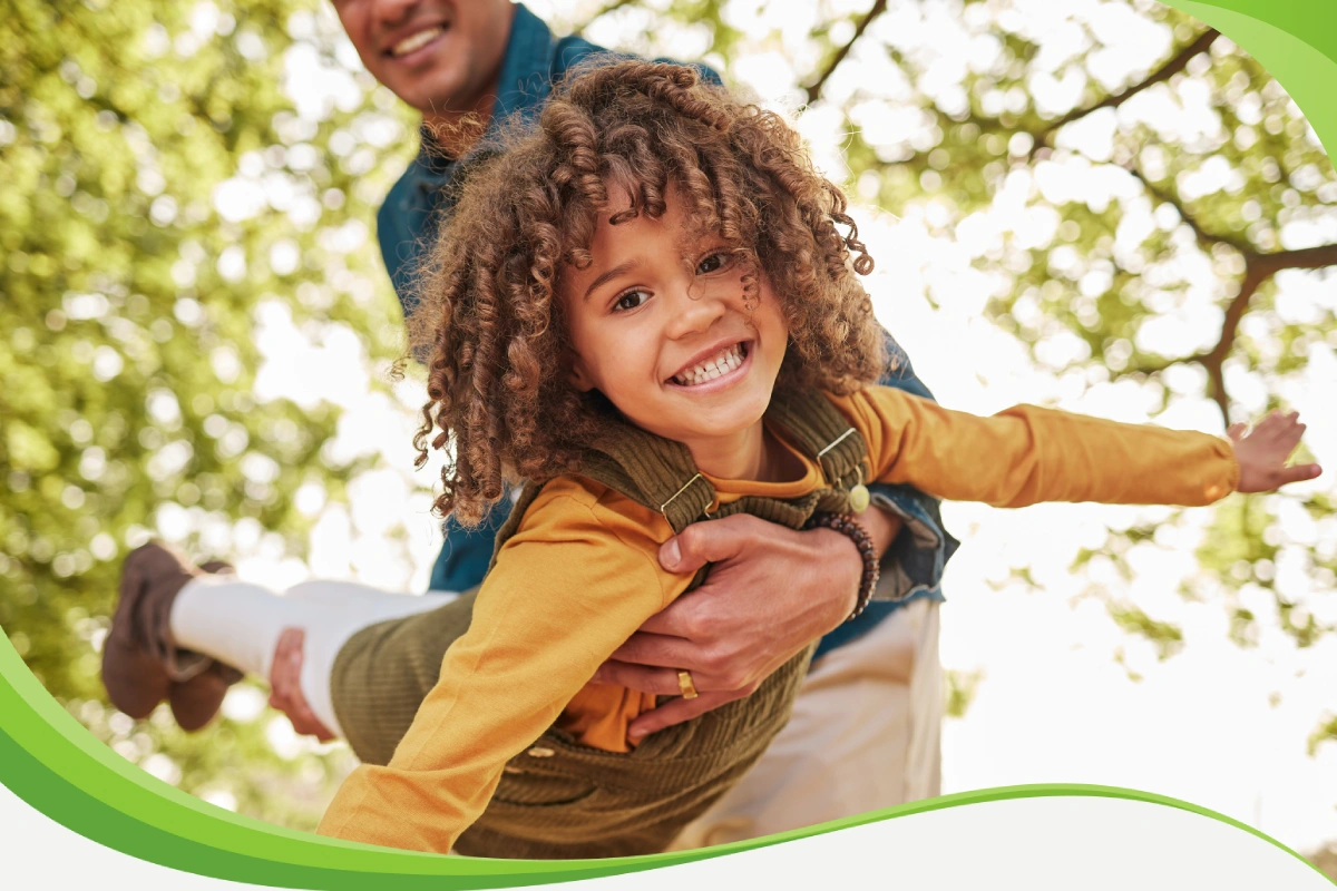 Menino sorridente, de braços abertos, içado pelo pai na horizontal, brinca como se pudesse voar.