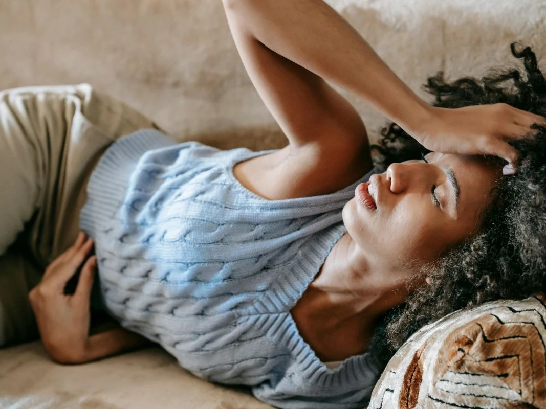 Mulher com expressão de dor sentada em um sofá, com uma mão na cabeça e outra na barriga, representando o mal-estar da dor de cabeça com enjoo.