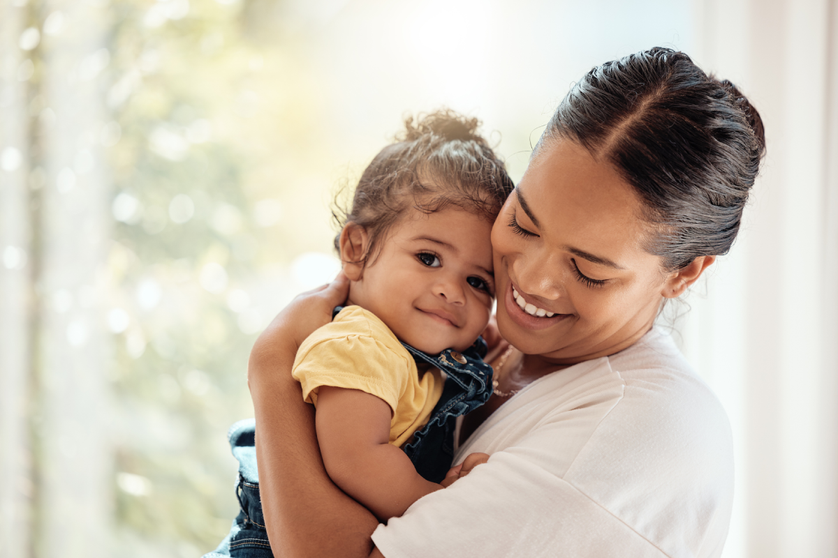 Mãe sorrindo e segurando menininha. A pequena está olhando para a câmera.