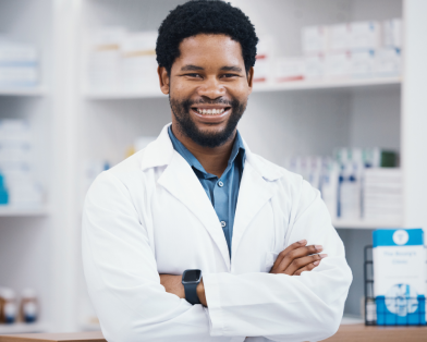 Farmacêutico sorridente de braços cruzados em uma farmácia, pronto para orientar sobre o uso correto de medicamentos.