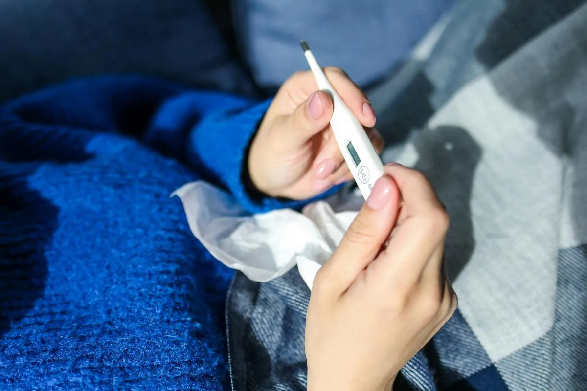 Close de mãos segurando um termômetro digital e um lenço de papel amassado, sugerindo a medição de temperatura durante um resfriado ou gripe.