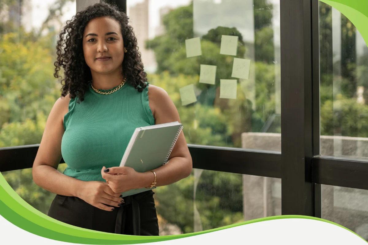 Mulher segurando uma pasta de trabalho diante de uma janela. Ao fundo é possível observar uma área verde. Ela tem uma expressão serena.