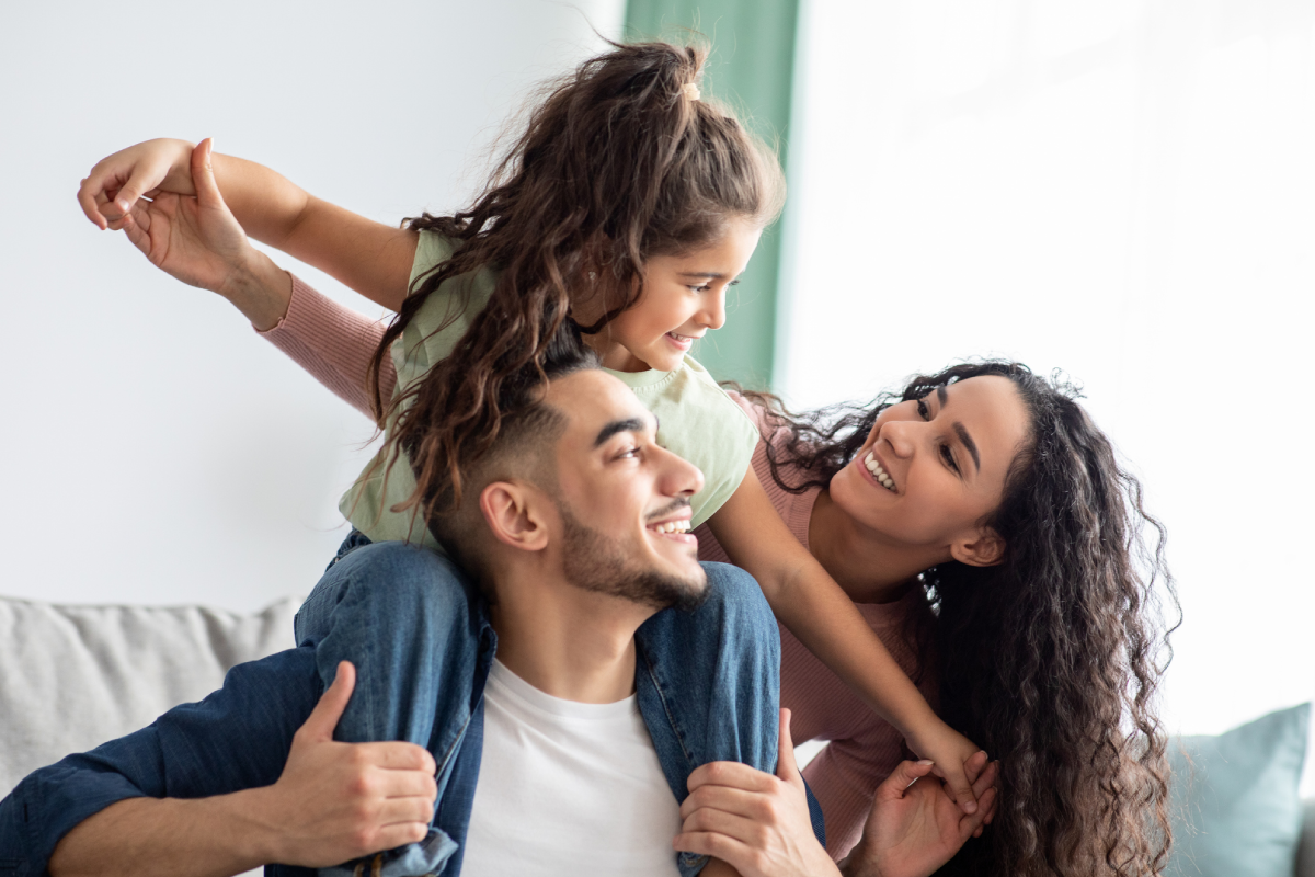 Família feliz no sofá. A filha está nas costas do pai, com os braços abertos, e a mãe olha para ela de forma afetuosa.