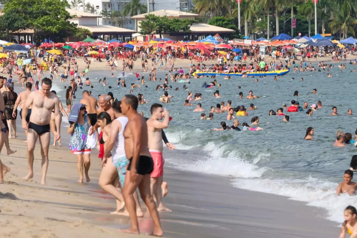 Uma praia lotada de pessoas aproveitando o mar e a areia em um dia ensolarado. Há guarda-sóis coloridos e banhistas tanto na beira quanto dentro da água, com prédios residenciais ao fundo cercados por vegetação tropical.