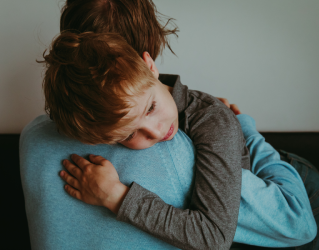 Garoto sendo acolhido por adulto, abraçado e com a cabeça em seu ombro, parece ter chorado.