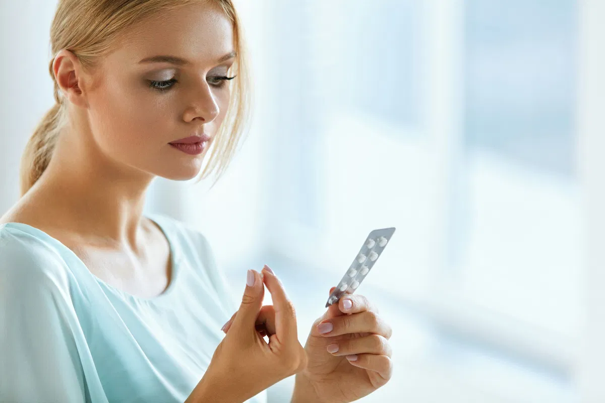 Uma mulher jovem, vestida com uma blusa azul clara, observa atentamente uma cartela de comprimidos enquanto segura um deles na outra mão. Está em um ambiente bem iluminado por luz natural, provavelmente em um cômodo tranquilo.