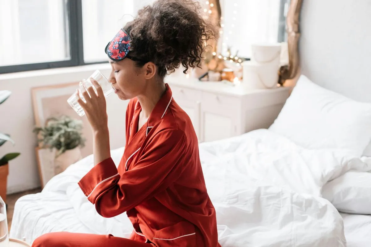 Mulher com cabelos cacheados e uma máscara de dormir floral, vestindo um pijama de seda vermelho, sentada na cama e bebendo um copo de água.